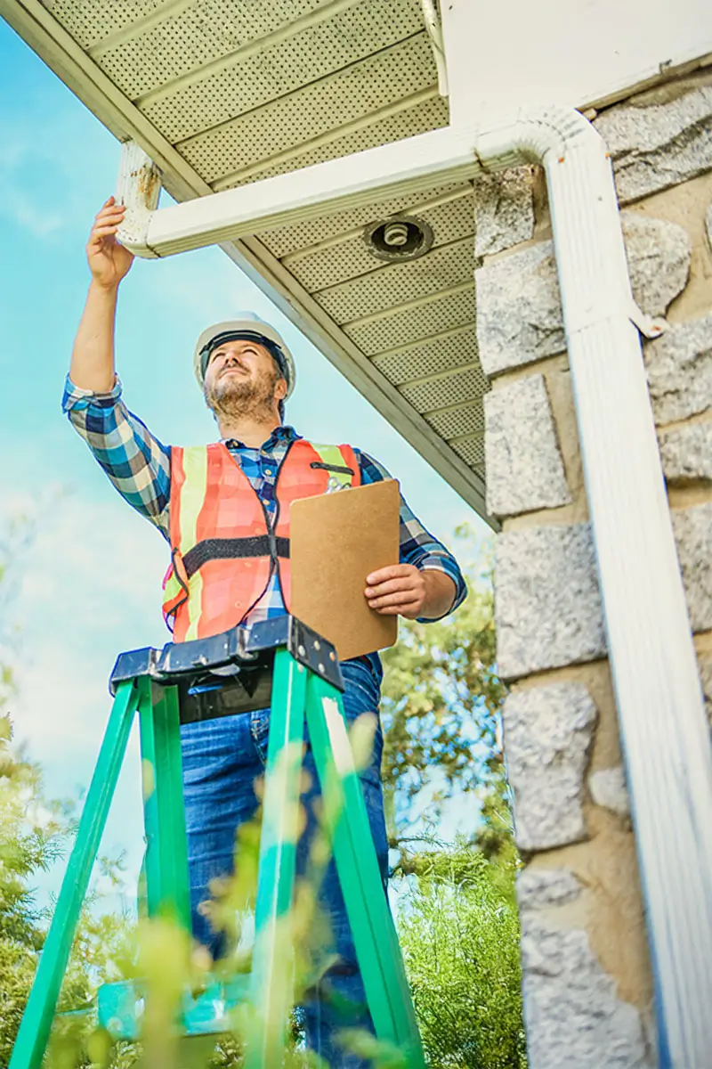 Man with hard hat standing on steps inspecting house gutter.